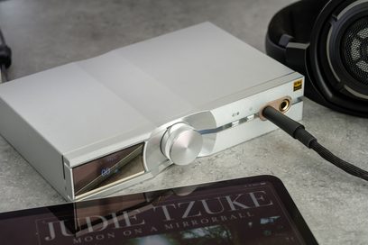 iFi Audio IDSD NEO sølvfarvet DAC-audiokonverter, set forfra, tilsluttet et sort hovedsæt med jackstik