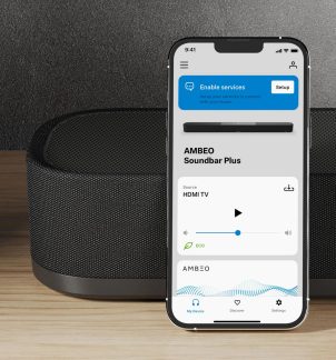 Chytrý telefon s rozsvíceným displejem a otevřenou aplikací, kovovými hranami, položený před černou reproduktorovou skříní s tkaným potahem, na dřevěném povrchu