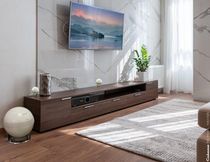 Moderne stue set fra trekvart vinkel med lavt, rektangulært tv-møbel i brun valnødfinish med skuffer og centralt rum, placeret på lyst trægulv foran marmormur med vægmonteret tv, cremefarvet tæppe og enkelte dekorative genstande