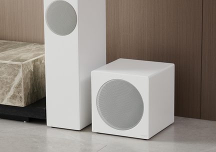 Bílý krychlový subwoofer, čelní panel s kruhovou kovovou mřížkou, pohled ze tří čtvrtin, u bílé reprosoustavy