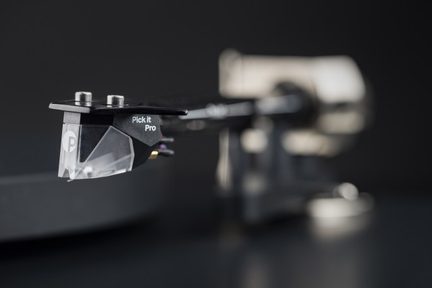 Celulă phono Pick it Pro neagră cu vârf transparent, braț de citire metalic neclar în fundal, fundal întunecat.