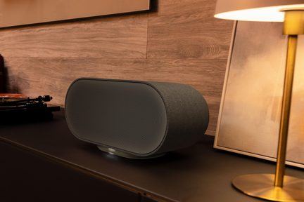 Sort oval trådløs Denon-højttaler med fin frontgrill, stående på tv-møbel i moderne stue med varm belysning
