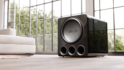Sort subwoofer med tre cirkulære åbninger, synligt logo, placeret på trægulv, lys sofa og store vinduer