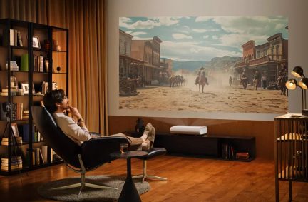 Muž sedící na černém křesle, který se dívá na projekci westernového filmu na stěně, vlevo police s knihami, vpravo stojací lampa