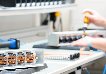 Šedý hliníkový obdélníkový kryt, pohled z tří čtvrtin zepředu, obsahující čtyři černé válcové moduly zarovnané za viditelnými měděnými svorkami, položený na světlém pracovním stole
