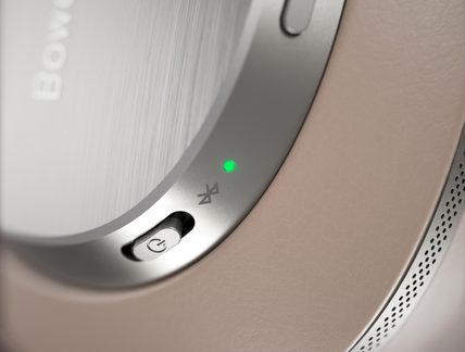 Nærbillede af en høretelefon-øreklokke i børstet metal og beige læder, tænd/sluk-skyder, Bluetooth-symbol og grøn LED