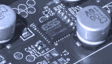 Nærbilde av kretskort, svart ESS-brikke, sylindriske metallkondensatorer, hvite markeringer, elektroniske komponenter