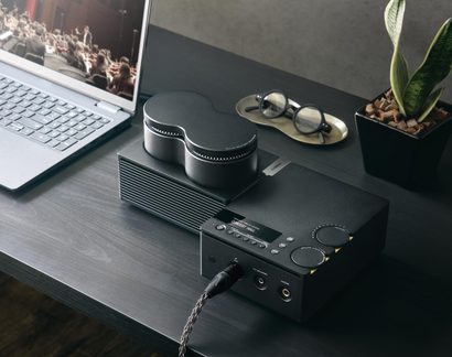 Amplificatore audio da scrivania nero in metallo, frontale con display, manopole, due jack e cavo intrecciato, vista dall'alto su una scrivania