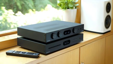 Deux appareils audio noirs empilés avec boutons et écrans, télécommande noire à gauche, enceinte blanche à droite, plante verte derrière