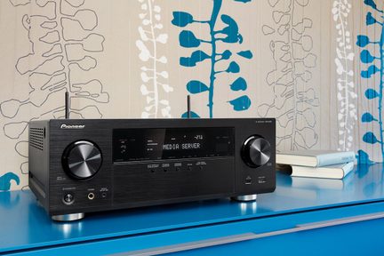 Sort audio-/videoreceiver på blå hylde, set forfra, knapper og stik synlige, to cylindriske genstande foran