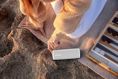 Bílý bezdrátový reproduktor Sonos Roam SL položený na písku, držený v ruce ženy v béžovém svetru, vedle hry backgammon
