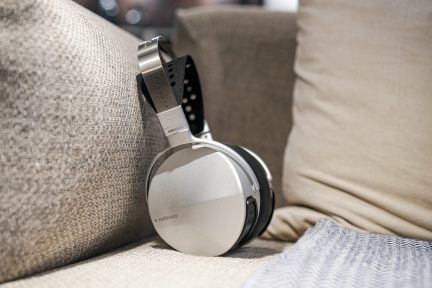 Sølvfarvede hovedtelefoner med sort perforeret hovedbøjle og sorte ørepuder, placeret på en beige stofsofa med et gråt plaid.