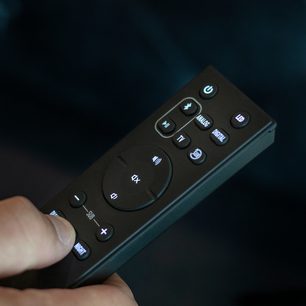 Sort Klipsch Cinema 400 fjernbetjening med blå baggrundsbelyste knapper, tæt forfra-perspektiv på mørkeblå baggrund