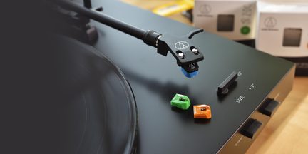 Sort vinylplade, skråt set, metal tonearm med blå pickup, to ekstra nåle i grøn og orange