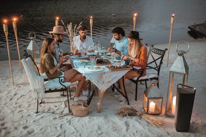 Billedet viser en gruppe mennesker, der spiser middag sammen på en strand i tusmørket, siddende omkring et bord, der er dækket med tændte stearinlys i lanterner