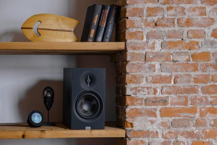 Wooden shelf, black speaker front view with woofer and tweeter, round screen and figurine, brick wall