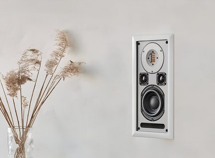 Altoparlante da parete incassato rettangolare bianco con cornice liscia, tre trasduttori neri ed apertura orizzontale, vista frontale, parete chiara, vaso in vetro trasparente a sinistra con steli di graminacee essiccate