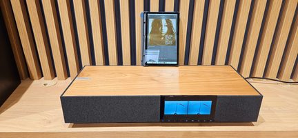 Rektangulært lydsystem set forfra let oppefra, kabinet i lyst træ med mørk kant, front i sort stof med indbygget skærm med dobbelt blå vu-meter og en række knapper, sort tablet indsat lodret i en bageste slids