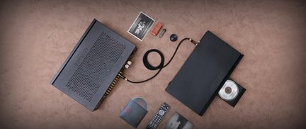 Sada dvou hi-fi audio zařízení ADVANCE v matné černé hliníkové barvě, pohled shora, levý kryt s kruhovými větracími otvory a konektory RCA, pravý kryt s otevřeným zásuvkou na CD obsahující disk, propojení černým audio kabelem