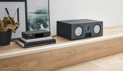 Boxă centrală hi-fi Canton neagră, cu amplificator de rețea compact asortat, pe un raft din lemn deschis la culoare