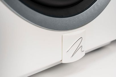 Subwoofer Dynamo 10 blanco lacado, vista cercana de una esquina inferior con logotipo metálico sobre superficie blanca y borde circular gris