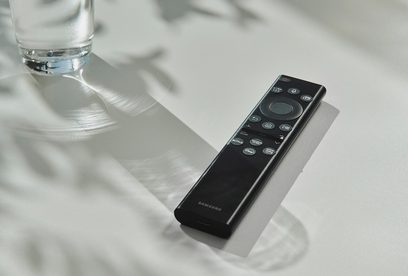 Černá dálková ovladač Samsung zepředu na bílém lesklém povrchu vedle průhledné sklenice vody