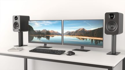 Arbeitsplatz von vorne gesehen mit zwei nebeneinander stehenden flachen, grauen Monitoren mit schmalem Rahmen, einer schwarzen Kunststoff-Tastatur und -Maus, zwei rechteckigen schwarzen Lautsprechern mit Doppelchassis auf Metallständern, alles auf einem weißen Schreibtisch platziert