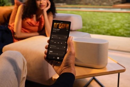 Hvid trådløs højttaler, der styres via smartphone, i moderne stue med kvinde liggende på sofaen