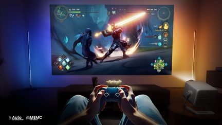 Vista subjetiva de um jogador segurando um controle azul e preto de plástico brilhante, em frente a uma grande projeção colorida de videogame em uma parede escura, com um projetor compacto cinza metálico posicionado lateralmente e duas barras de luz verticais emitindo halos azulados e alaranjados
