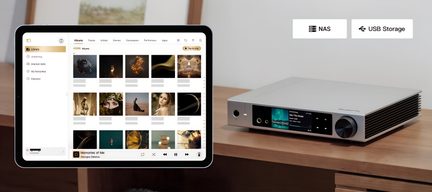 Dreiviertel-Frontansicht eines silbernen Hi-Fi-Audioplayers aus gebürstetem Aluminium mit großem schwarzem Drehknopf, Frontfarbdisplay und seitlichen Lüftungsgittern, auf einem Holzmöbel platziert, neben einem schwarzen Tablet im Querformat, das eine Musikbibliothek anzeigt