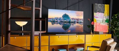 Samsung QE55Q85R-fladskærm, der viser et billede af isbjærge spejlet i vandet, stående på et gult møbel i et rum