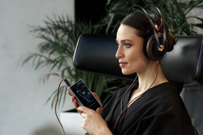 Siyah ve altın renkli Meze Audio 109 PRO kulaklık takan, elinde akıllı telefonu olan esmer kadın, yeşil bitkilerin önünde