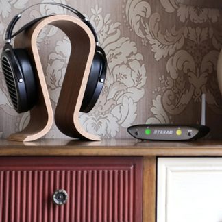 Forfra-vendt billede af et sort, circum-auralt headset med åbne ørekopper, placeret på en buet holder i lyst træ, ved siden af en lille sort lyd-boks med mørkegrå front, hvorpå der står "STREAM" og to indikatorlamper i grøn og gul