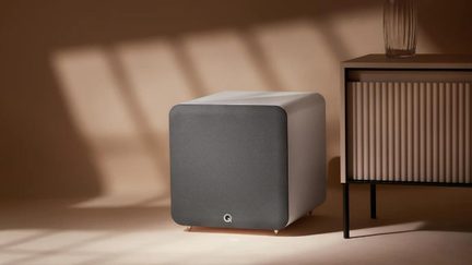 Matte white cubic subwoofer with rounded edges, dark gray fabric front grille featuring a small circular logo, standing on four metal feet, three-quarter right front view