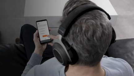 Πίσω όψη ενός ατόμου με μαύρα ακουστικά, γκρίζα μαλλιά, που κρατάει ένα smartphone με μια εφαρμογή στην οθόνη, καθισμένο σε μαύρο καναπέ.