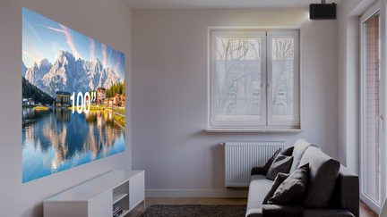 Salone luminoso, proiezione da 100" su parete bianca, proiettore nero a soffitto, divano grigio in tessuto, mobile TV bianco