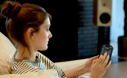 Frau auf einem Sofa mit Smartphone, das eine Audio-Steuerungs-App anzeigt, und Smart-Lautsprecher im Hintergrund