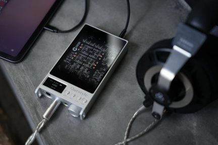 Silver Questyle M15 portable DAC headphone amplifier, connected to a smartphone and black headphones