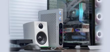 Compatto case audio in alluminio grigio, griglia laterale esagonale, frontale nero dotato di due manopole retroilluminate blu, porte audio, schermo e piedistallo, vista tre quarti anteriore