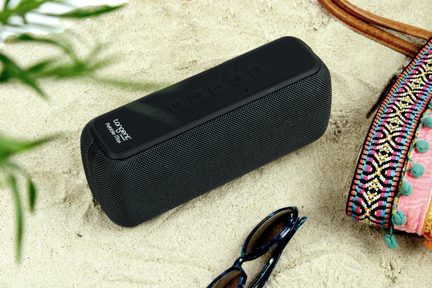Enceinte portable noire, grille en tissu, dessus en caoutchouc avec boutons en relief, vue en trois-quarts sur sable