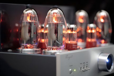 Pohled ze tří čtvrtin na lampový zesilovač, šedý hliníkový broušený kryt s velkým kulatým retro osvětleným modře tlačítkem, přední panel s zelenou LED a malými kovovými přepínači, horní část vybavená několika elektronkami v průhledném skle s oranžově žhnoucími vlákny