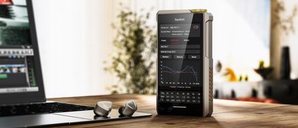 Reproductor de audio Hi-Fi FiiO de metal con pantalla y ecualizador, sobre escritorio de madera junto a un ordenador portátil y auriculares intrauditivos