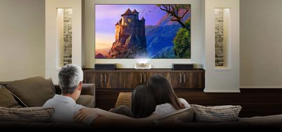 Benq W1600UST-videoprojektor, der projicerer et billede af et middelalderligt slot på en skærm i en stue med en sofa og to personer, der sidder med ryggen til