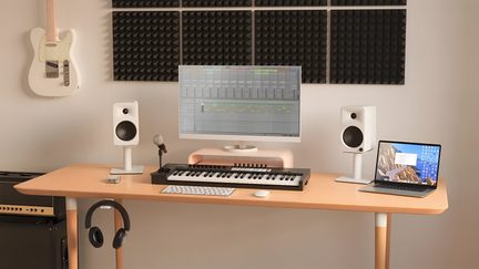 Heller Holztisch von vorne gesehen, ausgestattet mit zwei weißen Monitorlautsprechern auf Ständern, einem zentralen Computerbildschirm, einem schwarz-weißen Masterkeyboard, einem grauen Mikrofon auf Stativ, einer grauen Maus, einem geöffneten Laptop und einem aufgehängten schwarzen Kopfhörer, das Ganze vor schwarzen Akustikpaneelen platziert.