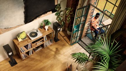 Ein Mann sitzt auf einem Balkon einer möblierten Wohnung mit Pflanzen, Regalen für Vinyl-Schallplatten und einem Holzcouchtisch