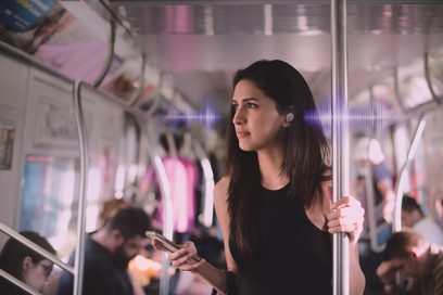 Ung brunette kvinde set fra siden i en overfyldt metro, iført hvide trådløse hovedtelefoner og holder en hvid smartphone