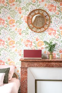 Triangle AIO 3 wireless speaker in Ochre Red placed on a marble furniture piece in front of vintage floral wallpaper