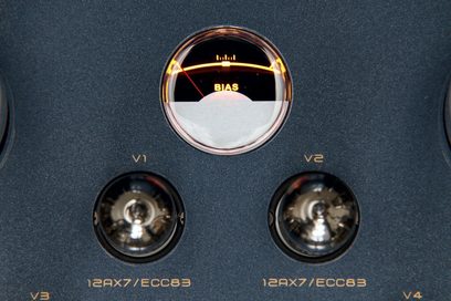 Detailní přední záběr na retro podsvícený kruhový měřič BIAS, obklopený dvěma lampami 12AX7/ECC83 v černém texturovaném kovovém šasi přístroje Cayin CS-55A KT88.