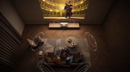 Oro vaizdas svetainės, ruda sofa, apvalus kavos staliukas, kilimas su geometriniais raštais, sieninis ekranas, projektorius, fotelis, užuolaidos.