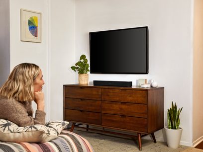 Trævogn til tv med en kvinde siddende og ser på et fjernsyn, en sort Sonos Beam-soundbar og en grøn plante